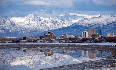 Anchorage, Alaska city buildings in distance with lake reflection during the daytime in winter. There are mountains behind the buildings